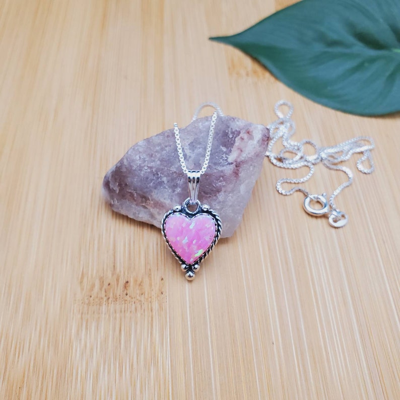 May include: A silver heart-shaped pendant necklace with a pink opal stone. The necklace is on a wooden surface with a small rock and a green leaf.