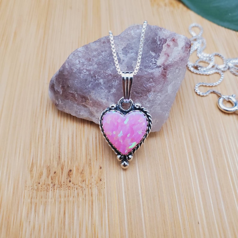 May include: A silver heart-shaped pendant necklace with a pink opal gemstone. The pendant has a delicate, ornate design and is set on a silver chain.