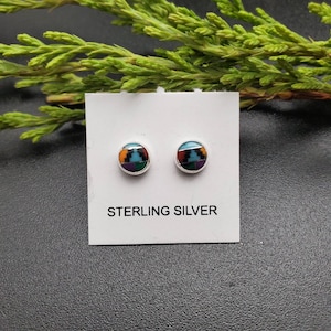 May include: A pair of round sterling silver stud earrings. Each earring features a mosaic design with orange, teal, black, purple, and red geometric shapes. The earrings are displayed on a white card with the words "STERLING SILVER".
