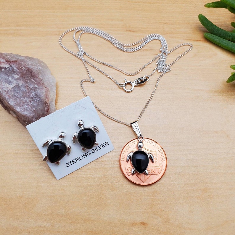 May include: Sterling silver necklace and earrings set featuring black onyx turtle pendants. The necklace is on a silver chain and the earrings are stud style. The set is displayed on a wooden surface with a penny for size reference.