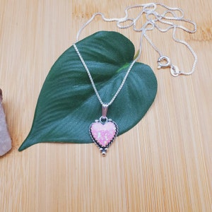 May include: A silver chain necklace with a pink opal heart pendant. The pendant has a detailed silver border and a small silver bead at the bottom.