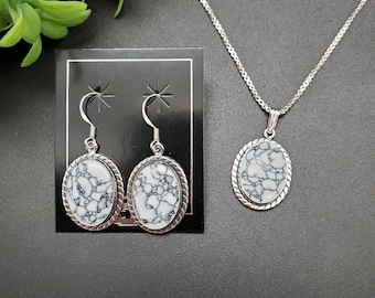May include: A silver necklace with a matching pendant and a pair of earrings. The oval-shaped pendant and earrings feature a white stone with black veining, set in a silver frame with a twisted rope design. The earrings are displayed on a black card.