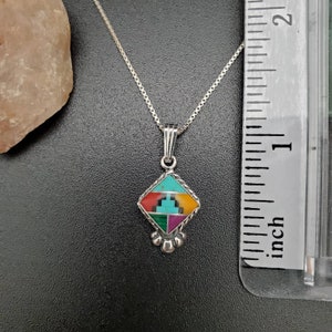 May include: A silver necklace with a diamond-shaped pendant featuring a multi-colored inlay of turquoise, red, green, and yellow stones. The pendant is approximately 1 inch in size.