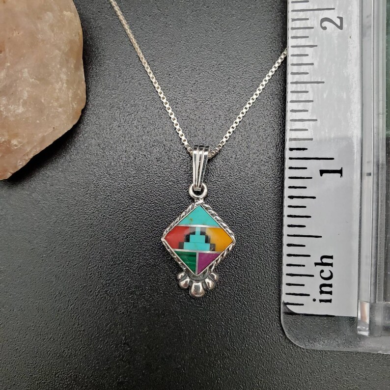 May include: A silver necklace with a diamond-shaped pendant featuring a multi-colored inlay of turquoise, red, green, and yellow stones. The pendant is approximately 1 inch in size.