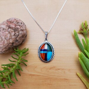 Pu&ograve; includere: Una collana con catena d'argento con un ciondolo ovale che presenta un design geometrico di pietre turchesi, rosse, verdi e blu incastonate.