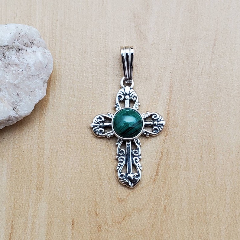 May include: A silver pendant in the shape of a cross, featuring an ornate design with a round, green malachite stone at the center. The cross has decorative flourishes and a bail for a chain. The pendant is displayed on a light wood surface.