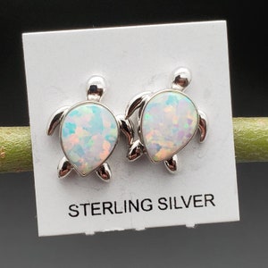 May include: A pair of sterling silver earrings featuring a turtle design with an iridescent opal stone in the center. The earrings are displayed on a white card with the text "STERLING SILVER".