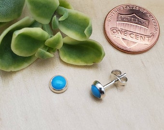 May include: A pair of turquoise stud earrings with a silver bezel setting. The earrings are round and have a bright blue stone. A penny is next to the earrings for size comparison. The earrings are on a light wood surface.