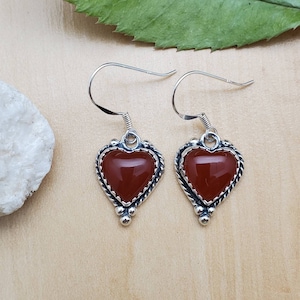May include: A pair of silver heart-shaped earrings with red carnelian stones. The earrings have a delicate, ornate design with small beads around the edges of the hearts.
