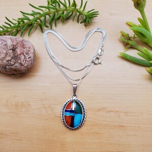 Pu&ograve; includere: Una collana con catena d'argento con un ciondolo ovale che presenta un design geometrico di pietre turchesi, rosse, arancioni, verdi e blu incastonate.
