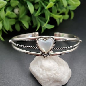 May include: A silver bracelet with a heart-shaped mother-of-pearl inlay. The bracelet has a twisted wire design and a double band.