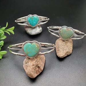 Op de afbeelding: Drie zilverkleurige manchetarmbanden, elk met een hartvormige turkoois steen. De turkoois stenen variëren in kleur van licht tot medium blauwgroen. De armbanden worden gepresenteerd op kleine, natuurlijke stenen tegen een donkere achtergrond.