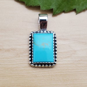 May include: A turquoise stone pendant with a silver frame. The pendant is rectangular and has a beaded border.