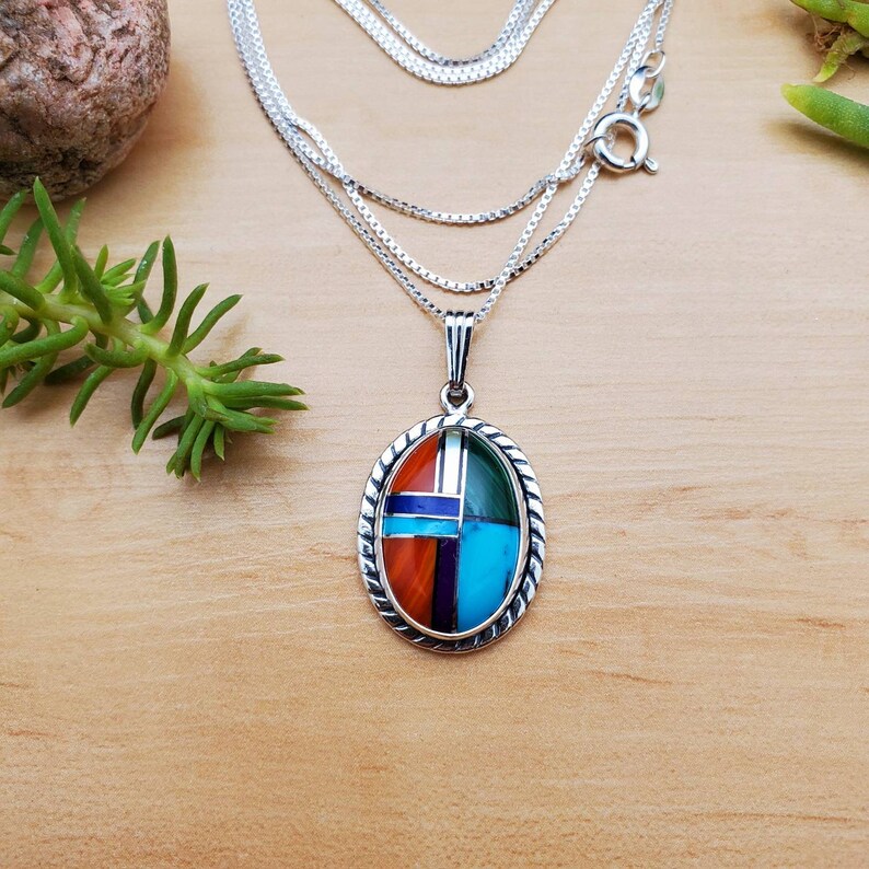 Pu&ograve; includere: Una collana con catena d'argento con un ciondolo ovale che presenta un design geometrico di pietre turchesi, rosse, verdi e blu.