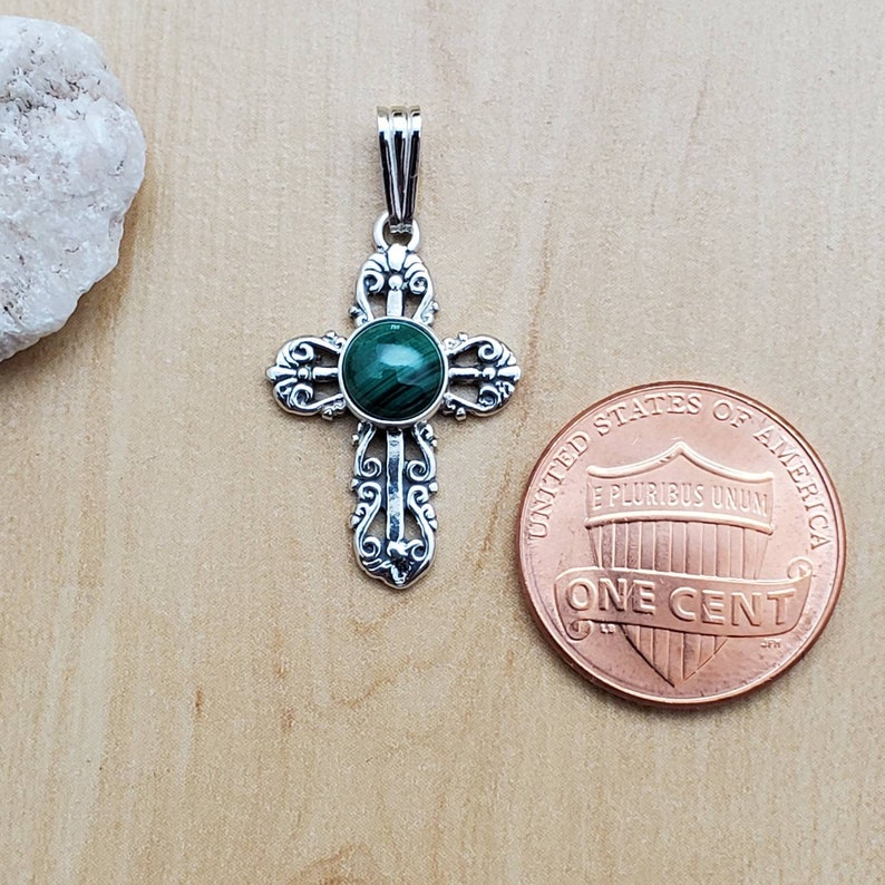 May include: A silver-colored pendant in the shape of a cross, featuring an ornate design and a round, green malachite stone at its center. A U.S. one-cent coin is next to the pendant for size comparison. The pendant is on a light brown surface.