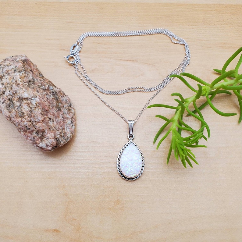 May include: A silver chain necklace with a teardrop-shaped opal pendant. The pendant has a silver border with a rope-like design.