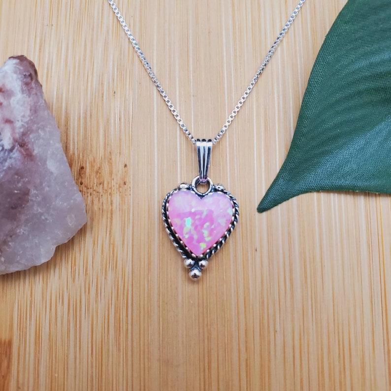 May include: A silver chain necklace with a pink opal heart pendant. The pendant has a detailed silver border and three small silver balls at the bottom.