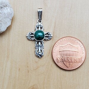 May include: A silver-colored pendant in the shape of a cross, featuring an ornate design with a round, green malachite stone at the center. The pendant is displayed next to a U.S. penny for size comparison.