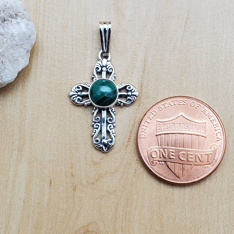 May include: A silver-colored pendant in the shape of a cross, featuring an ornate design with a round, green malachite stone at the center. The pendant is displayed next to a U.S. penny for size comparison.