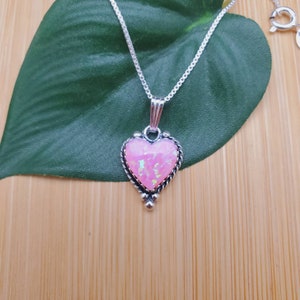 May include: A silver chain necklace with a pink opal heart pendant. The pendant has a detailed silver border and small silver beads at the bottom.