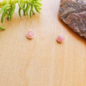 May include: A pair of small, round, pink gemstone earrings with silver settings. The earrings are on a light brown wooden surface.