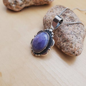 May include: A silver pendant with a purple amethyst stone set in a detailed silver frame. The pendant is on a silver chain.