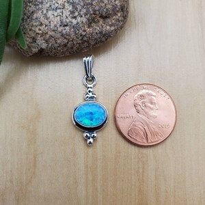 Puede incluir: Un colgante de plata con una piedra preciosa de &oacute;palo azul engastada en forma ovalada. El colgante tiene un aro con tres peque&ntilde;as cuentas a cada lado. El colgante se muestra junto a un centavo estadounidense para comparar el tama&ntilde;o.