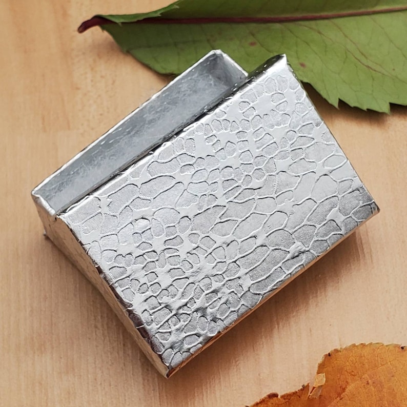 May include: A small, silver gift box with a textured, reptile-skin pattern. The box is open, revealing a silver interior. The box is angled, showing the inside. The background includes a wooden surface and a green leaf.