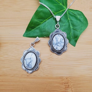 May include: Two silver pendants with white oval stones set in ornate silver frames. The pendants are on a wooden surface with a green leaf behind the top pendant.