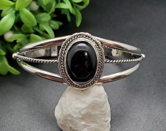 May include: A silver cuff bracelet with an oval black stone centerpiece. The stone is framed by a decorative rope-like border. The bracelet has a polished finish and a simple, elegant design.