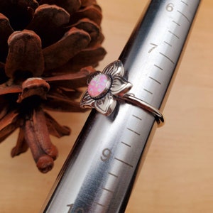 May include: A silver flower ring with a pink opal gemstone. The ring is sized on a metal ring sizer, showing a size 7.