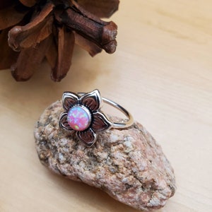May include: A silver ring with a pink opal flower design. The ring is set on a small rock.