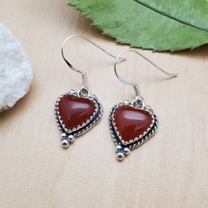 May include: A pair of silver heart-shaped earrings with red carnelian stones. The earrings have a delicate, ornate design with small beads around the edges.