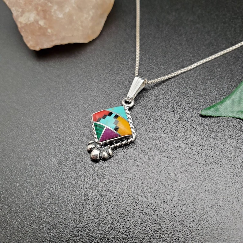 May include: A silver necklace with a geometric pendant featuring a colorful inlay of turquoise, red, green, purple, black, and yellow stones.