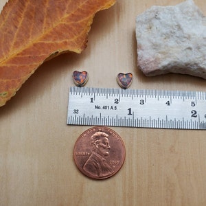 May include: Two silver heart-shaped earrings with a red, orange, and black opal stone set in each. The earrings are shown next to a ruler and a US penny for size reference.