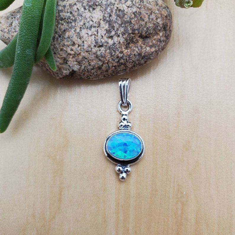 Puede incluir: Un colgante de plata con una piedra preciosa de &oacute;palo azul. El colgante tiene un dise&ntilde;o sencillo con una anilla en la parte superior y un peque&ntilde;o grupo de cuentas en la parte inferior.