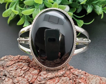 Puede incluir: Una pulsera de plata con una gran piedra ovalada negra. La pulsera tiene un diseño trenzado en la banda.