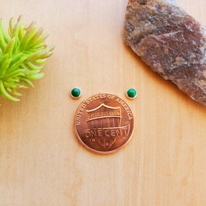 May include: A pair of small green gemstone earrings, shown next to a US penny for size comparison.