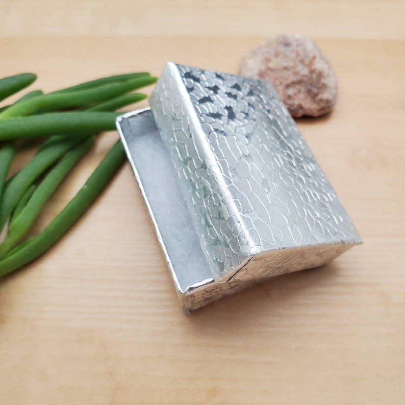 May include: A silver gift box with a textured, patterned surface. The box is open and the inside is lined with white paper.