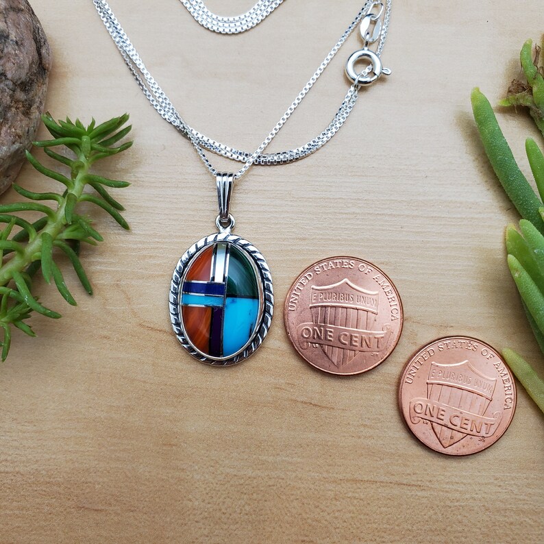 Pu&ograve; includere: Una collana con catena d'argento con un ciondolo ovale che presenta un design geometrico di pietre turchesi, rosse, verdi e blu incastonate. Il ciondolo &egrave; circa delle dimensioni di una moneta da un centesimo di euro.