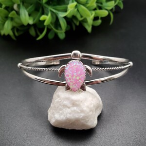 May include: A silver-toned cuff bracelet featuring a pink opal turtle design. The bracelet has a double-band design with a twisted rope detail. The turtle has a pear-shaped body and small flipper details. The bracelet is displayed on a white stone.