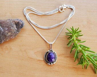 May include: A silver chain necklace with a purple gemstone pendant. The pendant is oval shaped and set in a silver frame with a floral design.