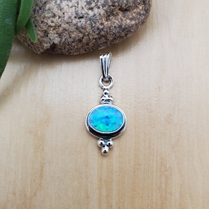 Puede incluir: Un colgante de plata con una piedra preciosa de &oacute;palo azul. El colgante tiene un dise&ntilde;o delicado con peque&ntilde;as cuentas y un aro.