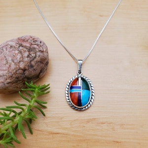 Pu&ograve; includere: Una collana con catena d'argento con un ciondolo ovale che presenta un design geometrico di pietre turchesi, rosse, verdi e blu incastonate.