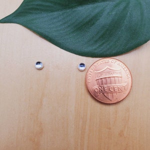 Peut inclure: Deux petites boucles d'oreilles en argent avec des pierres claires, à côté d'un centime d'euro pour la comparaison de taille.