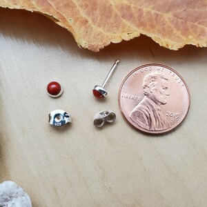 May include: A pair of silver earrings with red gemstone accents. The earrings are shown next to a US penny for size comparison. The penny has the inscription "In God We Trust" and "Liberty" on it. The year 2019 is also visible on the penny.