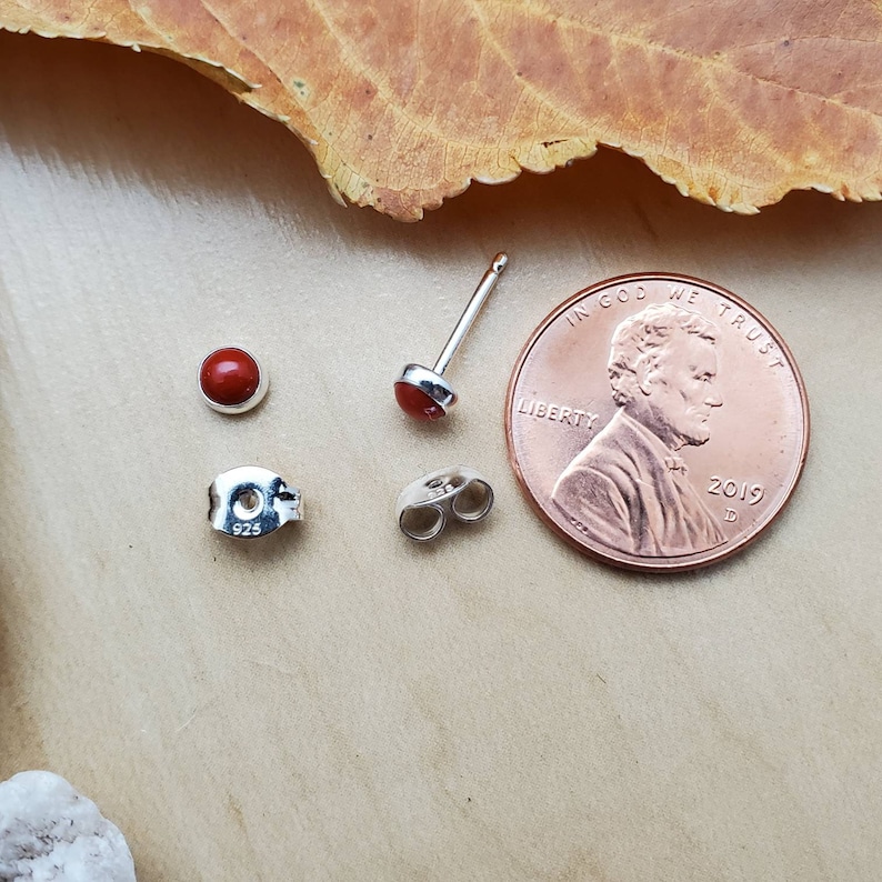 May include: A pair of silver earrings with red gemstone accents. The earrings are shown next to a US penny for size comparison. The penny has the inscription "In God We Trust" and "Liberty" on it. The year 2019 is also visible on the penny.