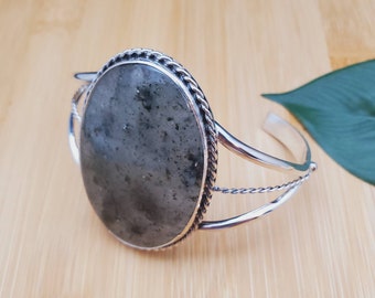 May include: A silver cuff bracelet featuring a large oval-shaped grey gemstone set in a rope-like border. The bracelet has a simple, elegant design.
