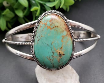 May include: A silver cuff bracelet featuring a large, oval turquoise stone with brown and tan veining. The stone is set in a decorative silver bezel with a rope-like border. The bracelet has a split shank design.