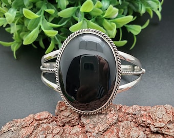 Puede incluir: Brazalete de plata con una gran piedra negra ovalada pulida. La piedra está engastada en un bisel de plata con un diseño de cuerda trenzada. El brazalete tiene un diseño de banda dividida.